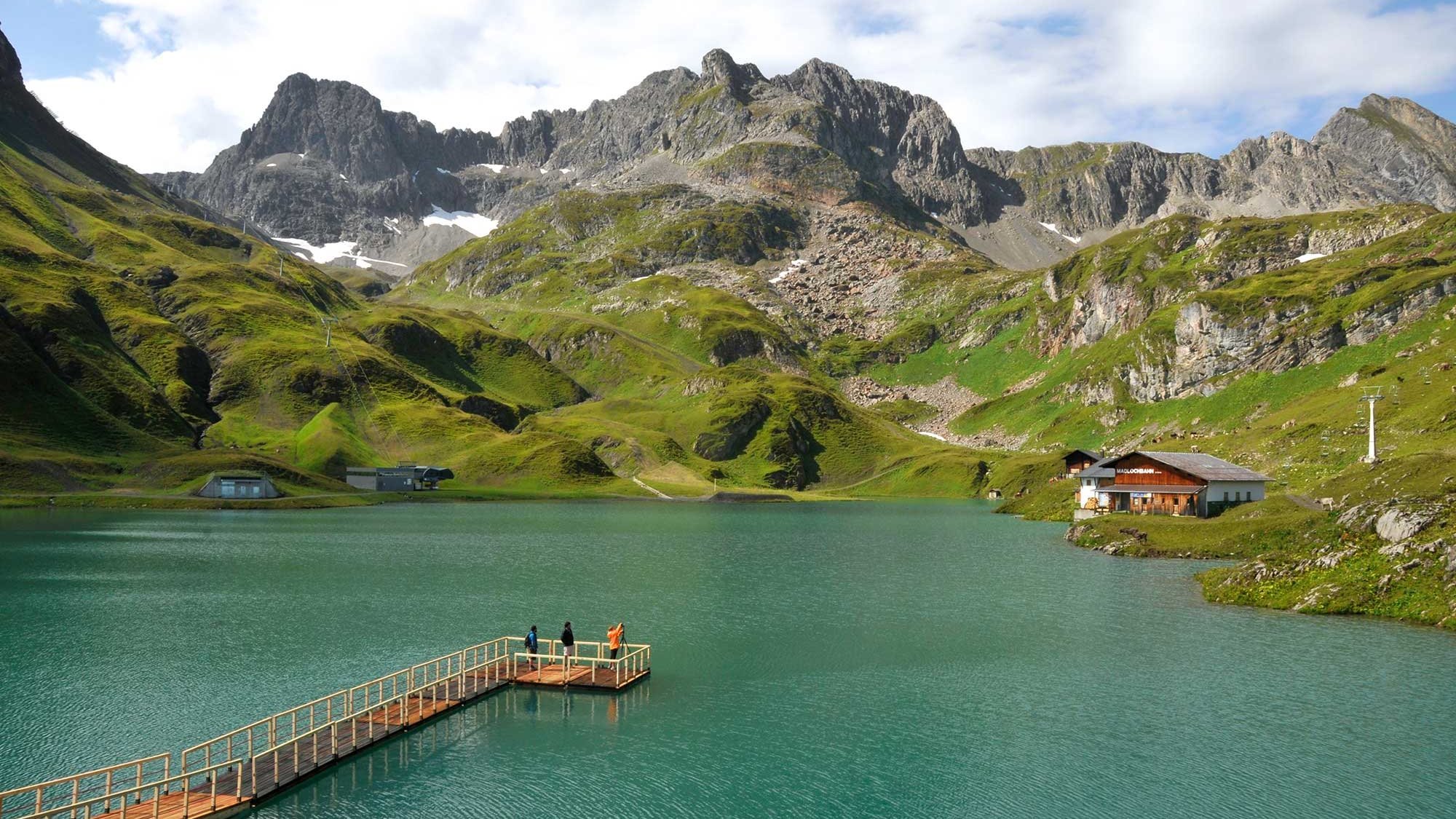 Bergsee mit Holzsteg und Menschen in alpiner Landschaft umgeben von Hügeln und Bergen