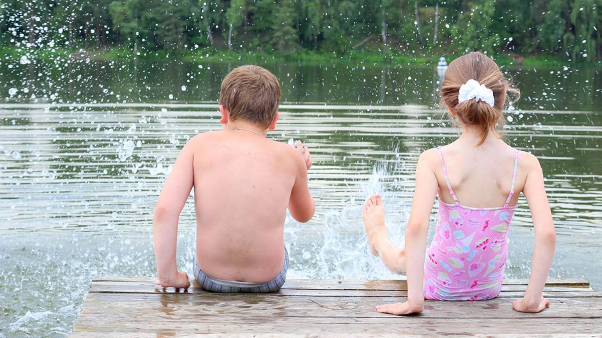 Zwei Kinder sitzen auf einem Holzsteg am See und planschen im Wasser während des Sommers.