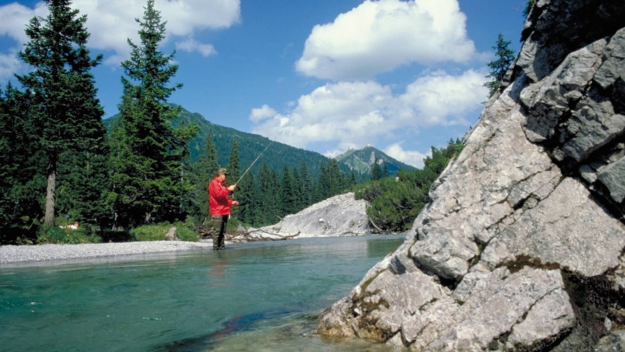 Eine Person fischt in einem klaren Fluss, umgeben von Bäumen und Bergen unter blauem Himmel.