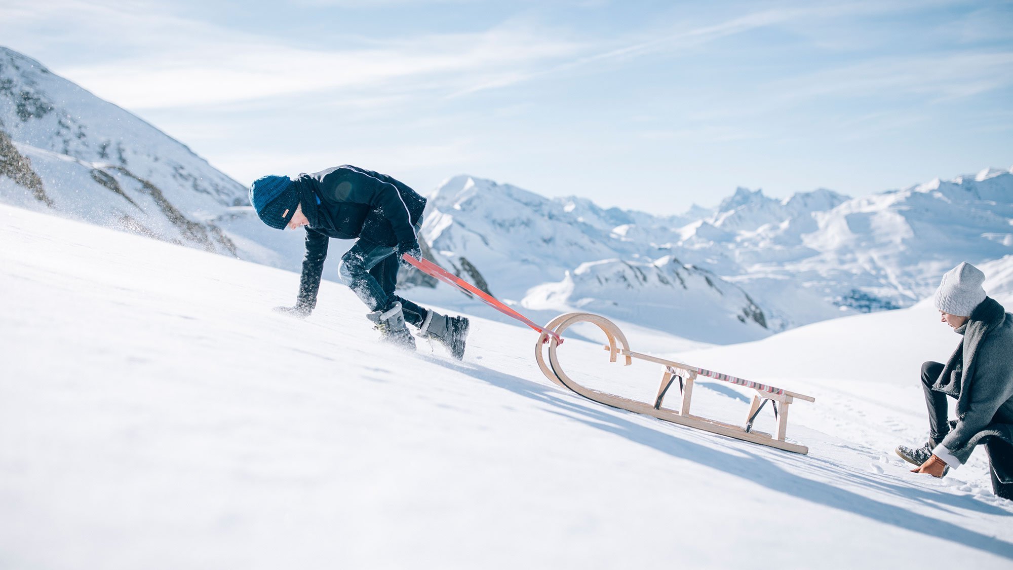 Zwei Personen im Schnee, eine zieht einen Schlitten, die andere sitzt daneben vor schneebedeckten Bergen