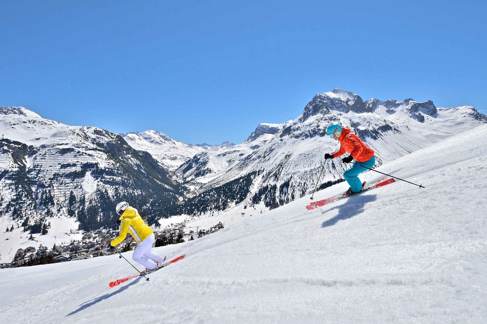 Zwei Skifahrer fahren eine verschneite Piste mit Bergen im Hintergrund bei klarem Himmel hinunter.