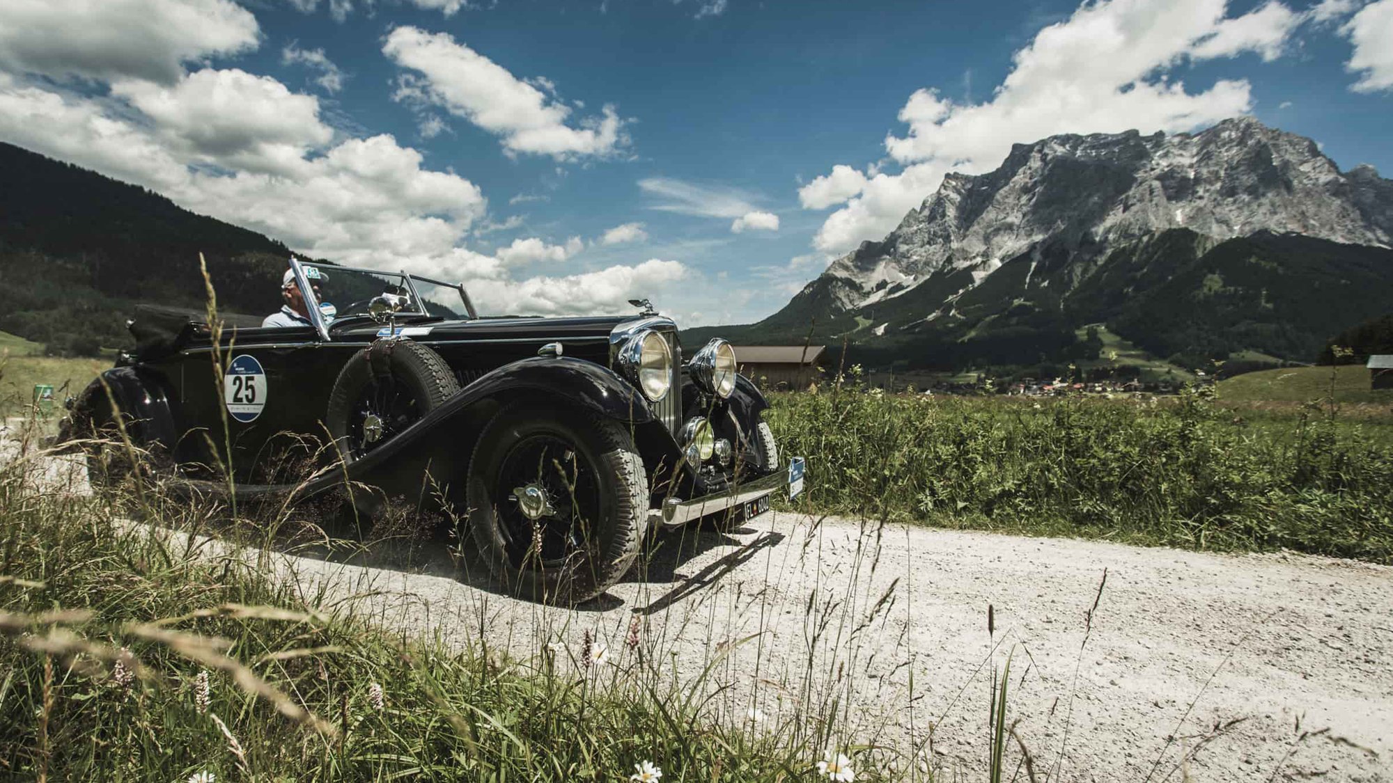Ein klassisches Automobil fährt auf einem Schotterweg durch eine Berglandschaft.