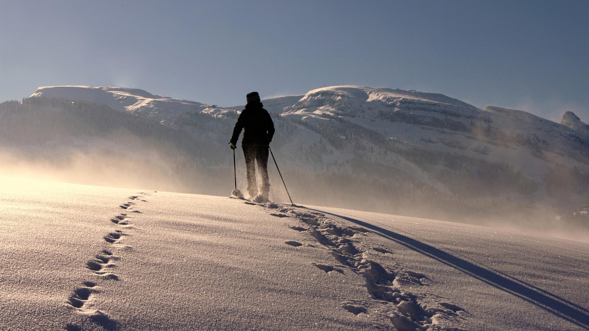 Person wandert mit Schneeschuhen durch eine verschneite Landschaft mit Bergen im Hintergrund