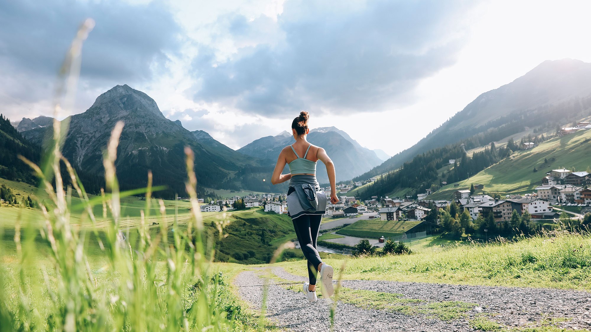 Eine Frau läuft auf einem Schotterweg durch eine gebirgige Landschaft mit einem Dorf im Hintergrund.