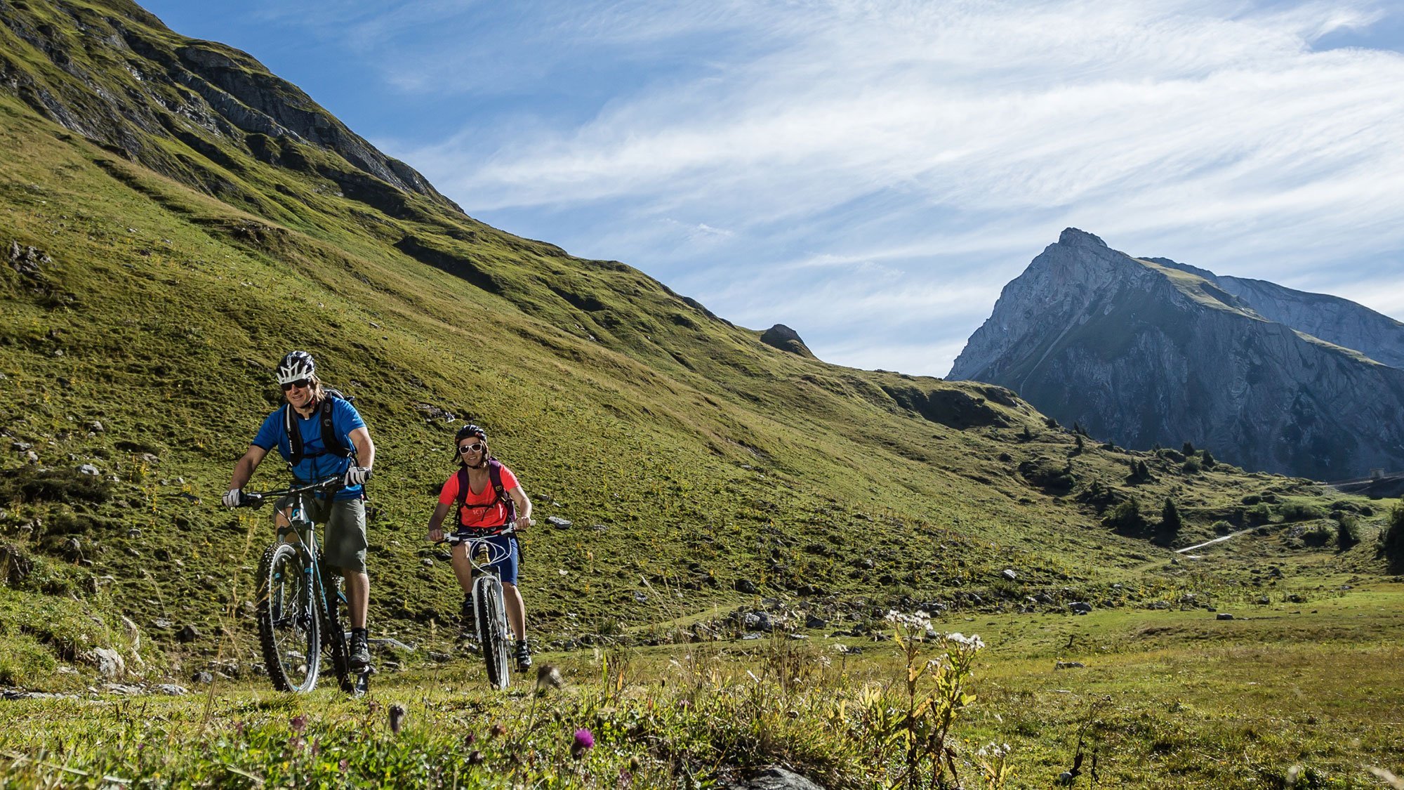 Zwei Radfahrer auf Mountainbikes fahren einen grasbewachsenen Weg durch die Berge.