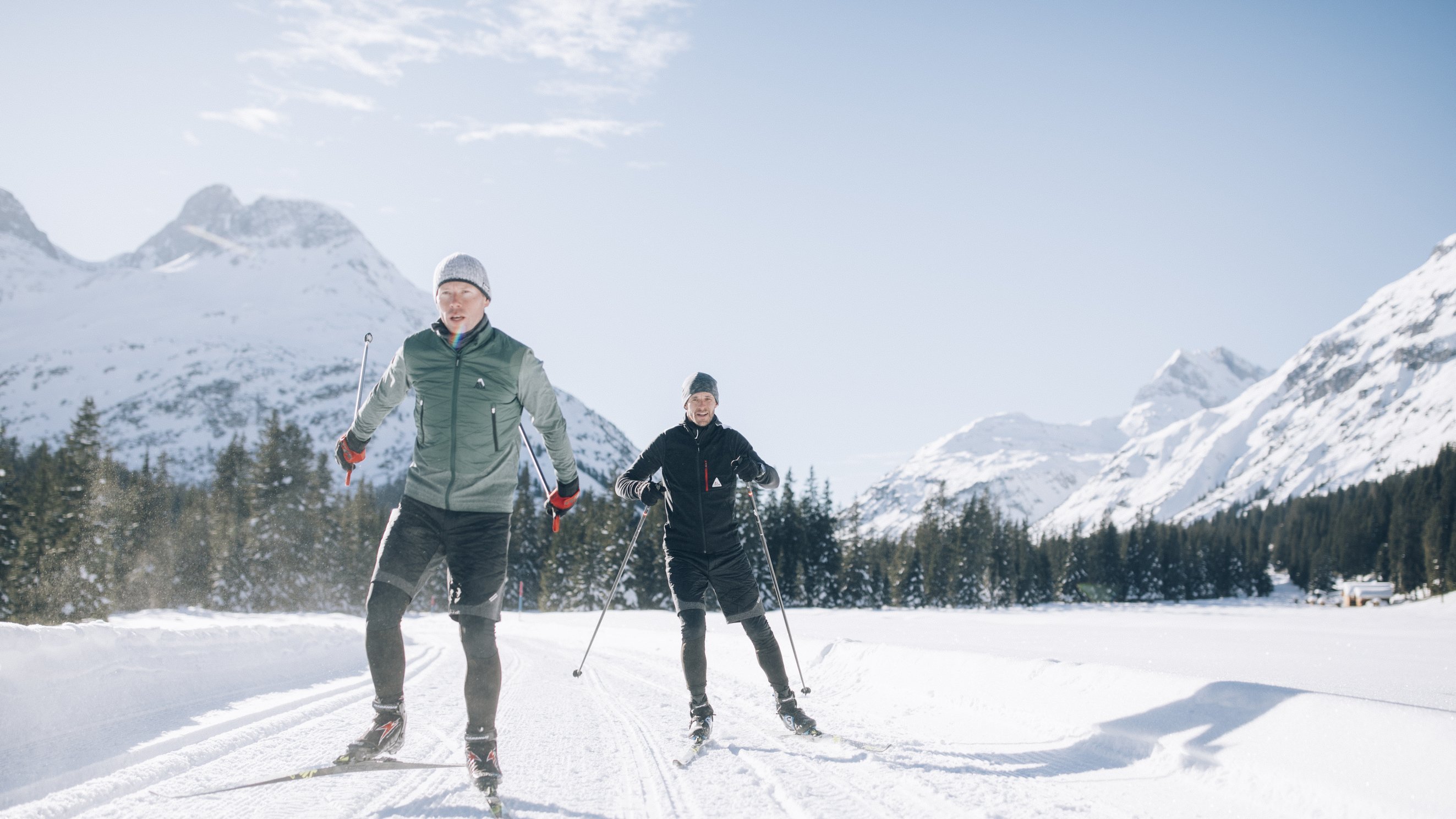 Zwei Personen beim Langlauf auf einem verschneiten Pfad mit Bergen und Bäumen im Hintergrund.