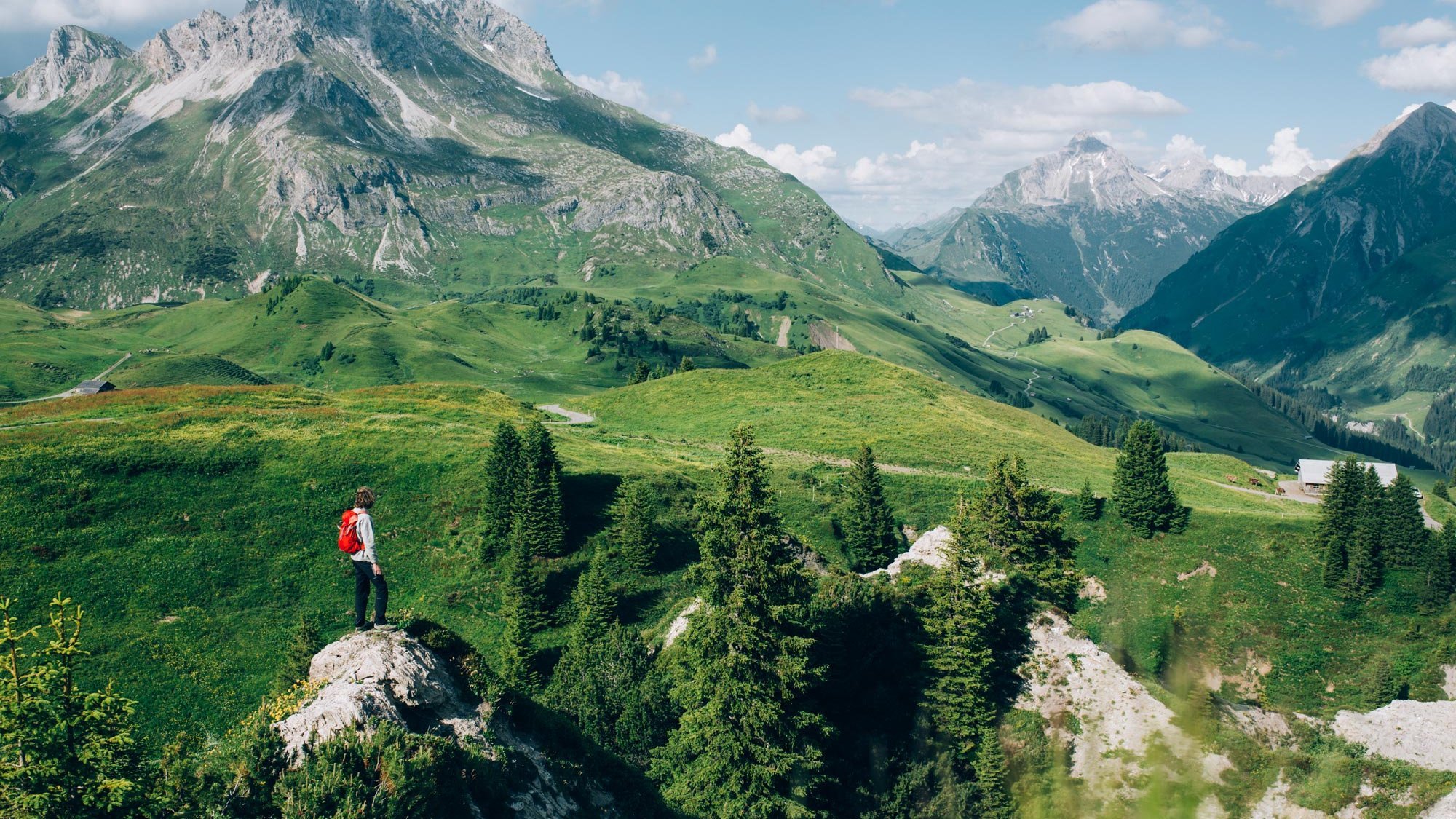 Person auf Felsen in einer grünen Berglandschaft umgeben von hohen Gipfeln und klarem Himmel