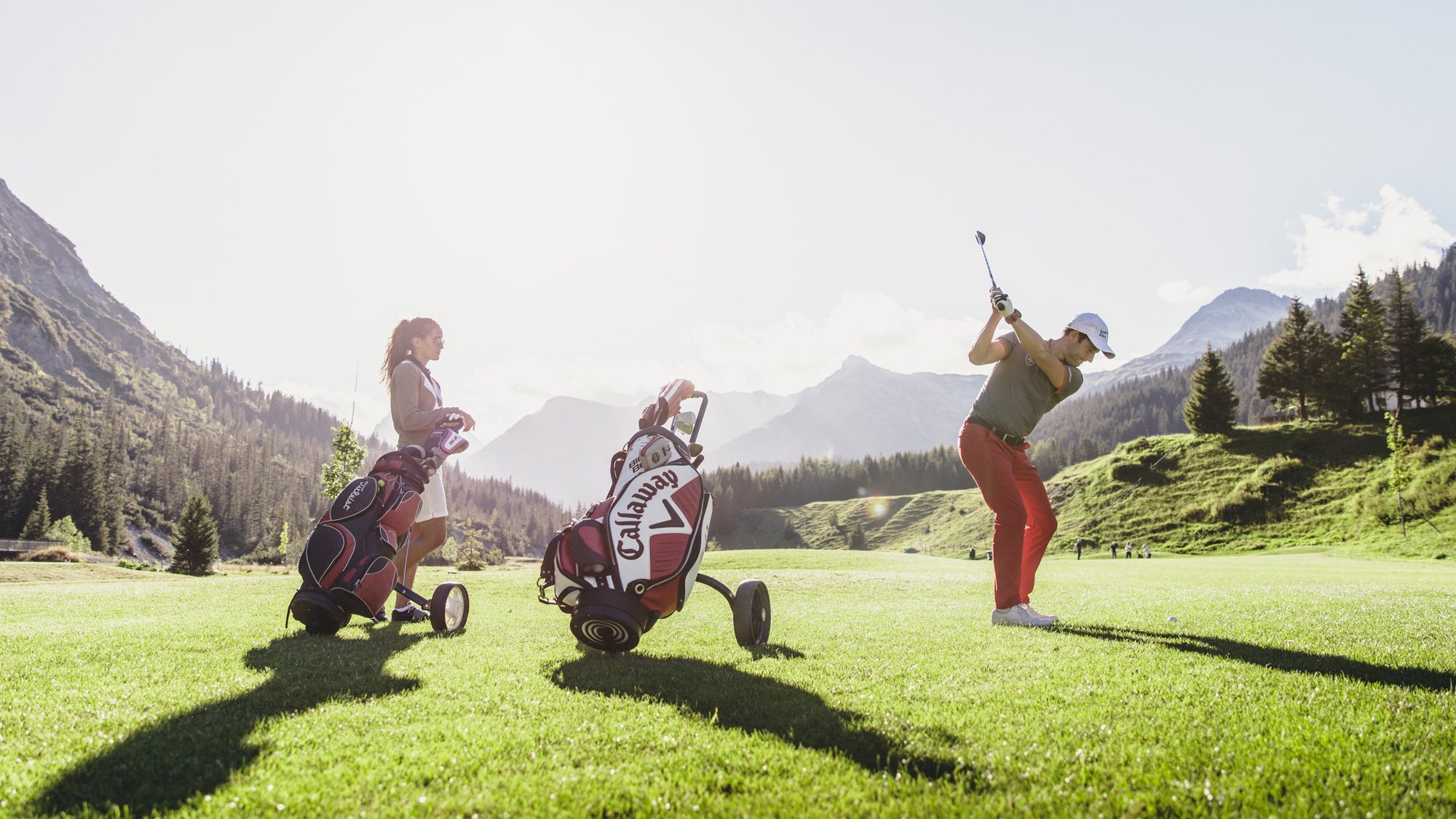 Zwei Frauen auf einem Golfplatz mit Golfausrüstung und Schläger in der Hand, eine bereit zum Abschlag.