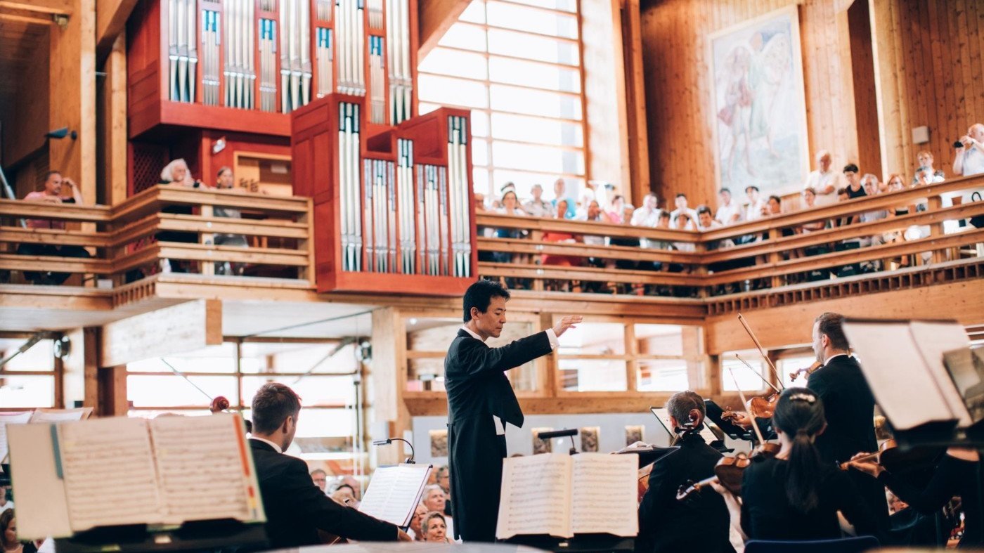 Dirigent leitet ein Orchester in einem voll besetzten Konzertsaal mit Publikum im Hintergrund.