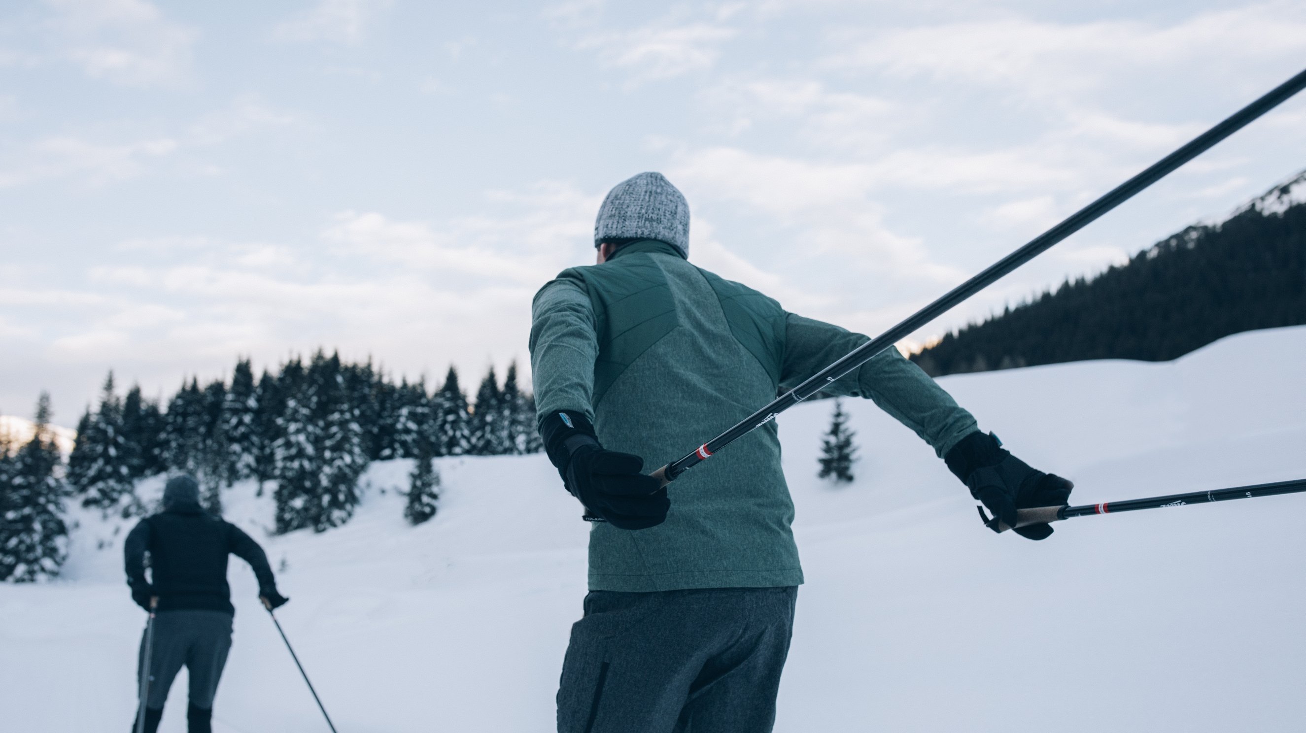 Zwei Personen beim Langlauf auf einer schneebedeckten Fläche mit Bäumen im Hintergrund und klarem Himmel.
