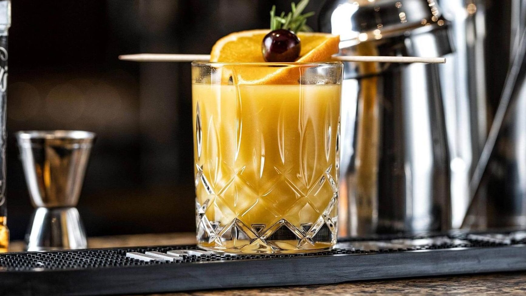 Cocktail in Kristallglas mit Orangenscheibe und Kirsche auf Bartresen neben Jigger und Shaker.