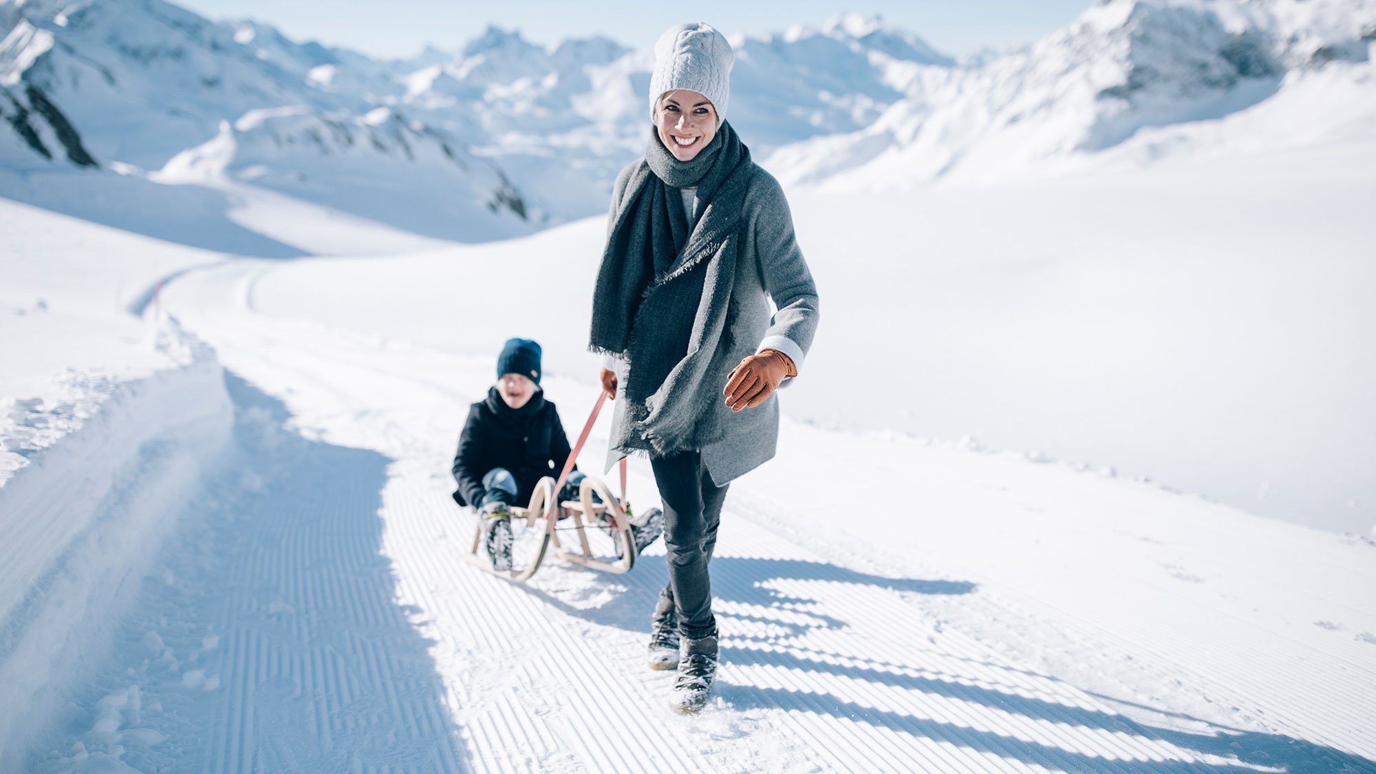 Eine Frau zieht ein Kind auf einem Schlitten durch eine verschneite Landschaft mit Bergen im Hintergrund.