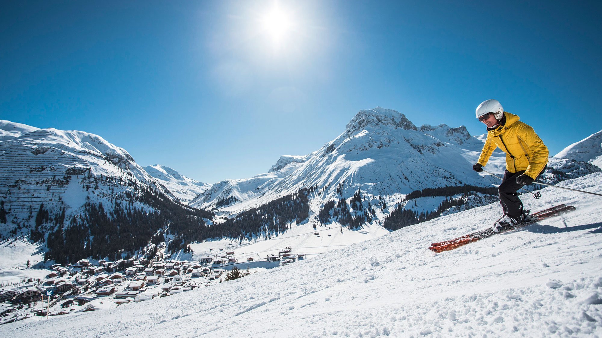 Skifahrer auf einer verschneiten Piste mit Bergen und einem Dorf im Hintergrund unter blauem Himmel