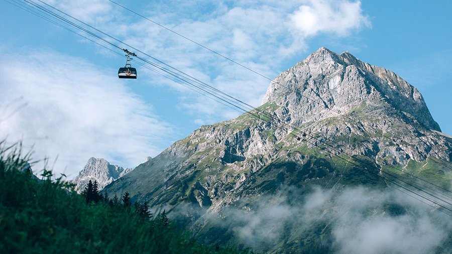 Eine Seilbahn fährt über grüne Landschaften und Wolken unter blauem Himmel auf einen Berg.