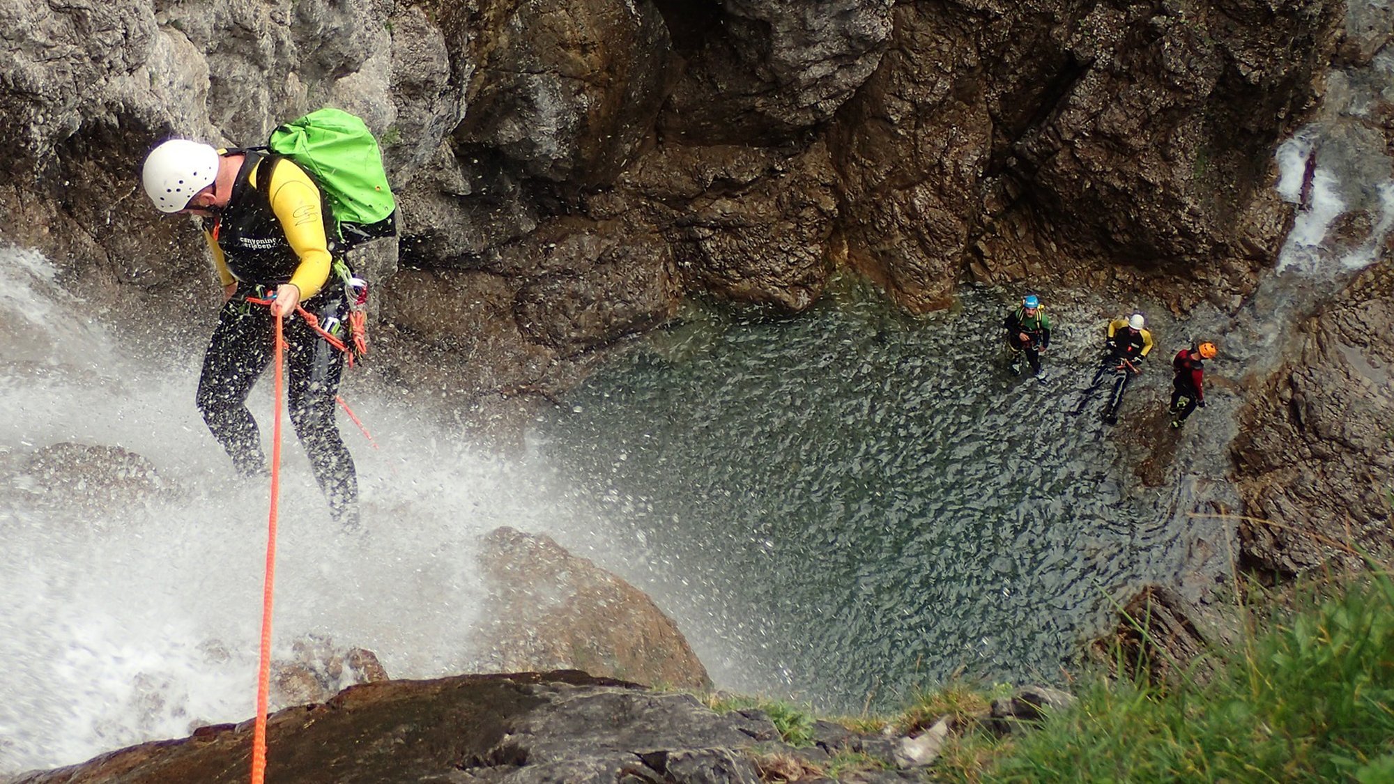 Person descendiert eine Felswand mit einem Seil, Wasserfall in der Nähe, Gruppe im Wasser.