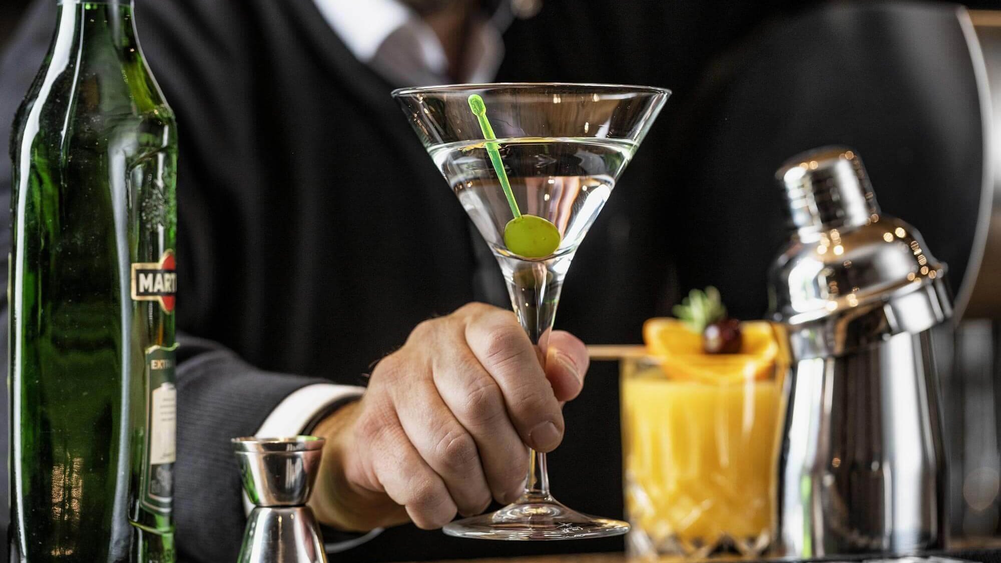 Bartender hält ein Martini-Glas mit einer grünen Olive, daneben Vermouth und ein Cocktailshaker.