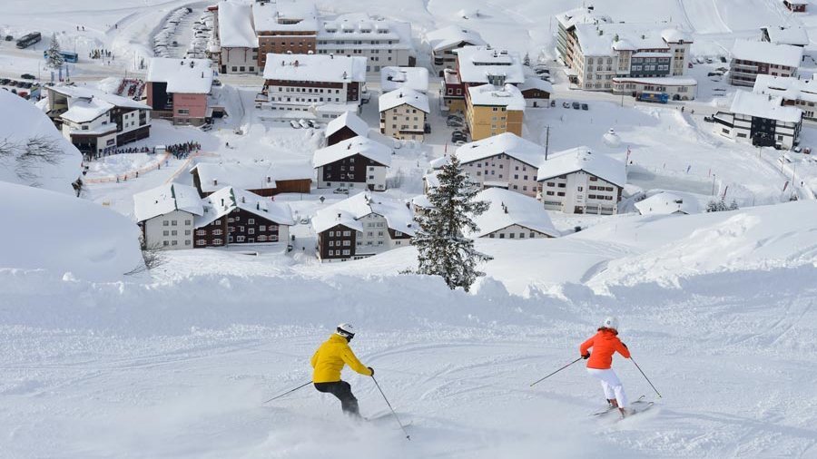 Zwei Skifahrer fahren eine verschneite Piste in einem malerischen Bergdorf hinunter.