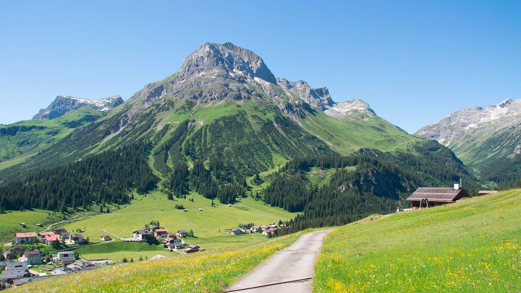 Berglandschaft mit einem gewundenen Pfad, umgeben von grünen Wiesen und Bergen, kleines Dorf im Tal