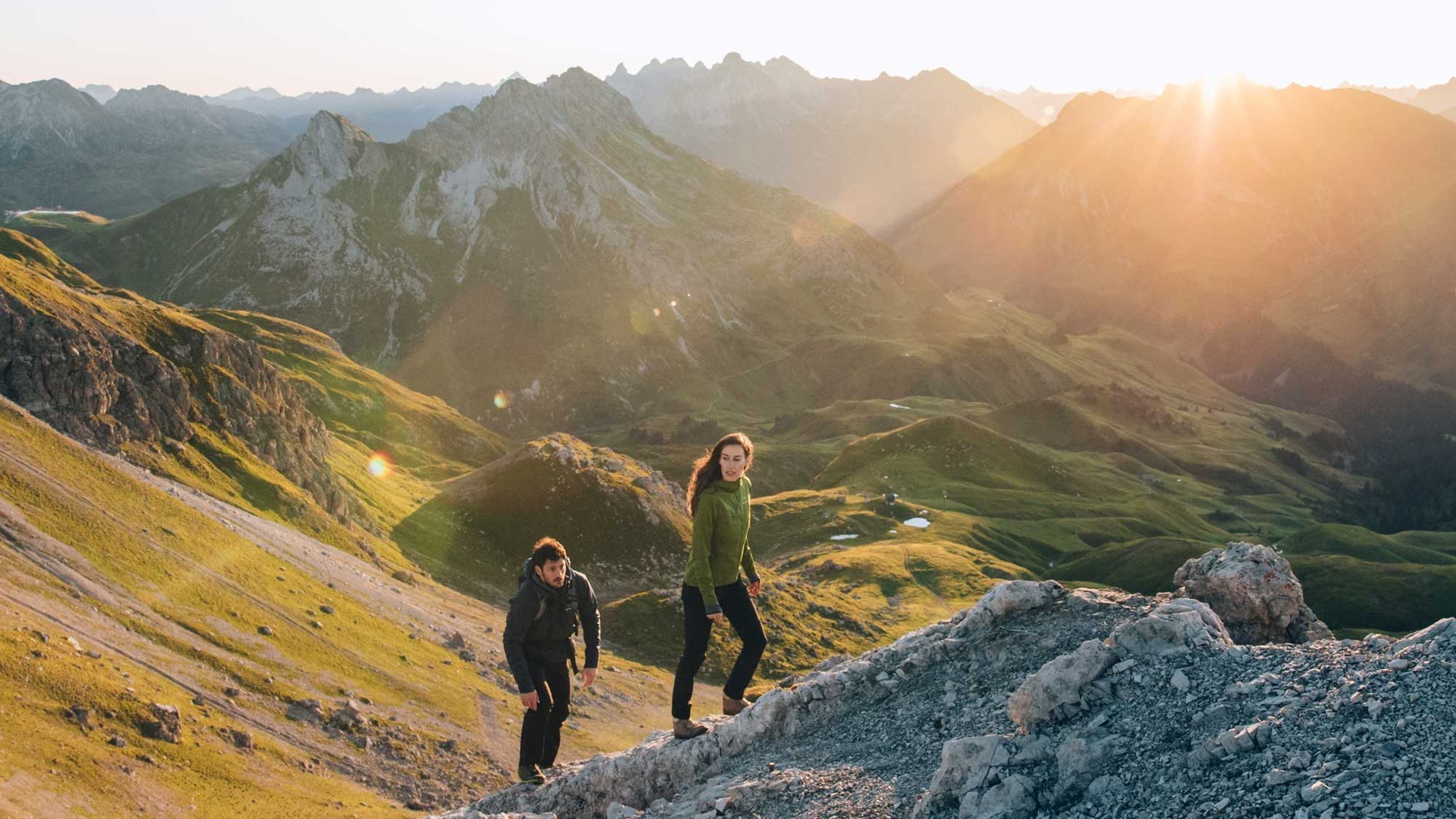 Zwei Wanderer erkunden ein felsiges Gelände vor einer beeindruckenden Bergkulisse bei Sonnenuntergang.