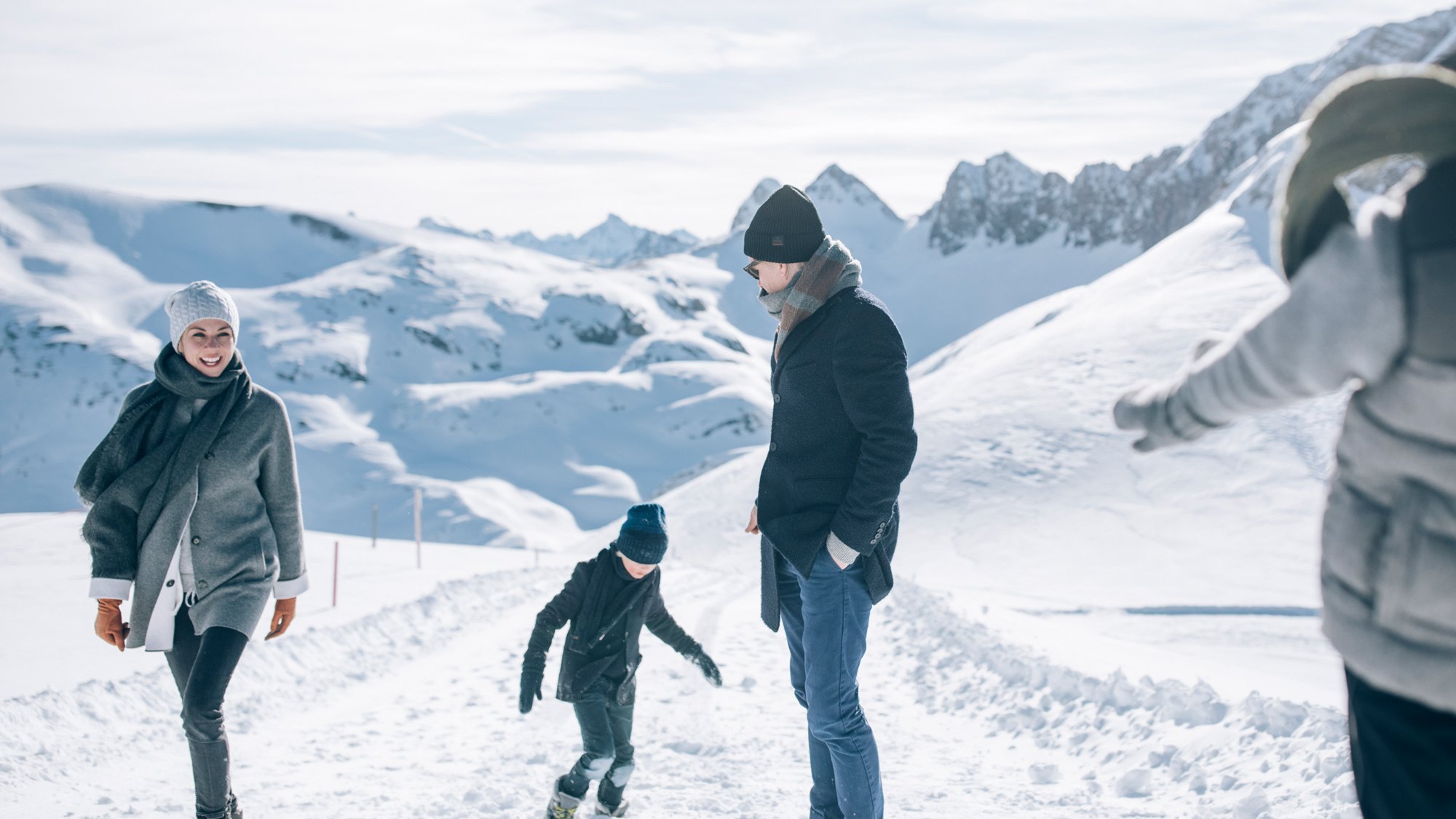 Familie wandert durch eine verschneite Landschaft mit Bergen im Hintergrund in Winterkleidung.