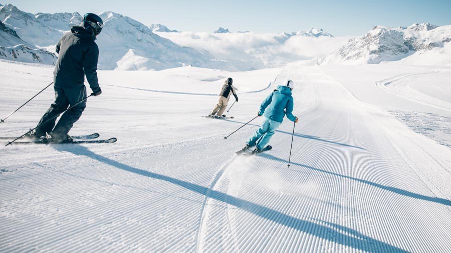 Drei Skifahrer fahren die schneebedeckte Piste mit Berglandschaft im Hintergrund hinunter.