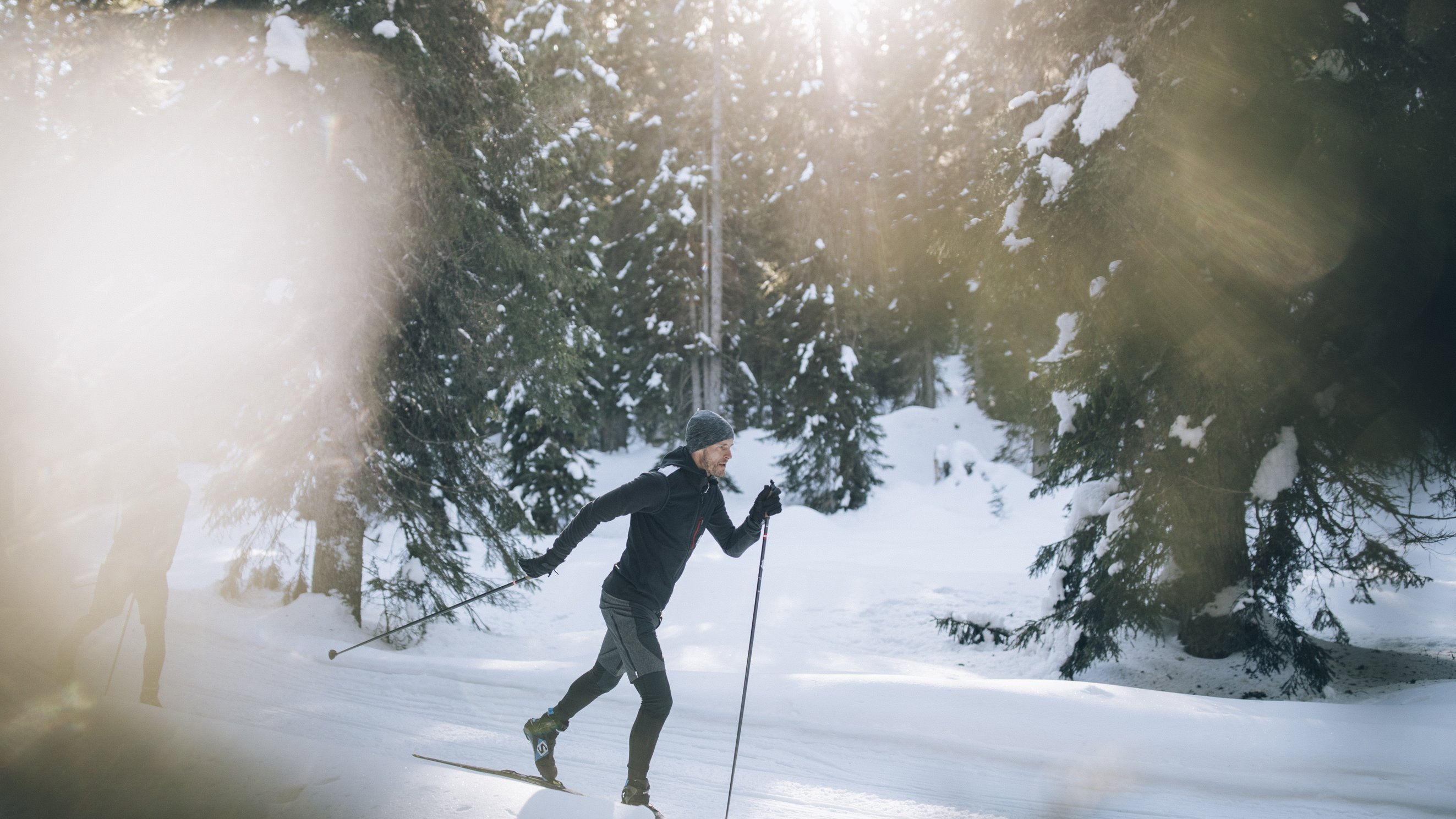 Eine Person fährt Langlauf durch einen verschneiten Wald mit Sonnenstrahlen, die durch die Bäume scheinen.