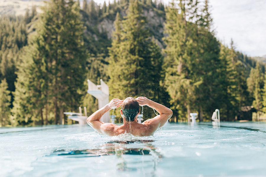 Person schwimmt in einem Außenpool mit Vorarlberger Berg- und Waldkulisse im Hintergrund © Daniel Zangerl